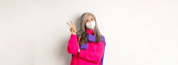 コヴィド パンデミック 社会的距離概念 コロナウイルスのマスクをした陽気でスタイリッシュなアジア系の先輩女性が勝利サインを見せ 白い背景の上に立って — ストック写真