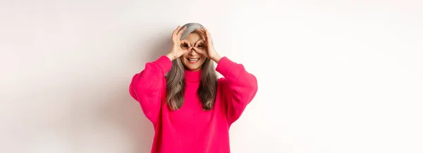 幸せと面白いアジアのシニア女性作る手双眼鏡 指と笑顔を通して見て 白い背景にピンクのセーターに立って — ストック写真