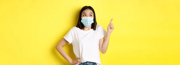 Covid Health Care Pandemic Concept 身穿医疗面罩和白色T恤的亚洲女性 手指指向左上角的标志 显示晋升 黄色背景 — 图库照片