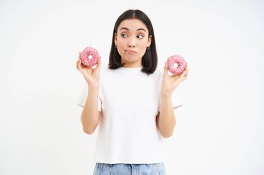 Sevimli Koreli kız, elinde iki tane pembe çörek tutuyor, düşünceli görünüyor, diyetini bozuyor, abur cubur yiyor, beyaz arka plan.
