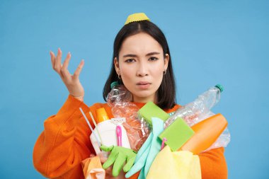 Geri dönüşüm kurallarıyla kafası karışmış kadın portresi, elinde boş plastik şişeler ve çöpler, karmaşık görünüyor, mavi arka plan.