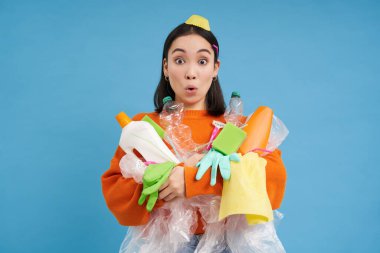 Şaşırmış yüzlü Koreli bir kadının portresi. Elinde boş plastik şişeler ve geri dönüşüm için çöp var. Mavi arka plan..