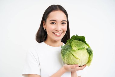 Genç Asyalı kadının yakın portresi, gülümsüyor ve gururla kendini yetiştirmiş sebzesini gösteriyor, elinde lahana, beyaz arka plan.