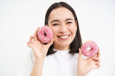 Gülümseyen Koreli kadın pembe jöleli donut yiyor, lezzetli çörekleri, abur cuburları seviyor, diyeti umursamıyor, beyaz arka plan.