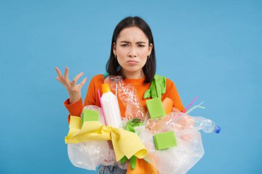 Kızgın genç kadın yumruğunu sıkar, sıralanmamış plastik atıkları tutar, geri dönüşüm yapar, sinirli görünür, mavi arka plan.