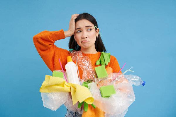 Женщина со сложным лицом, запутавшись в пластиковом мусоре, не знает, как перерабатывать предметы, зеленый, стоя на синем фоне изолированы.