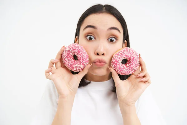 stock image Portrait of funny asian woman, tempted to eat tasty glazed dougnut, holding two donnuts and smiling excited, isolated on white background.