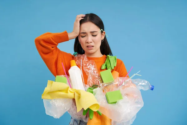 stock image Woman with complicated face, looking confused at plastic garbage, clueless how to recycle items, go green, standing over blue background isolated.