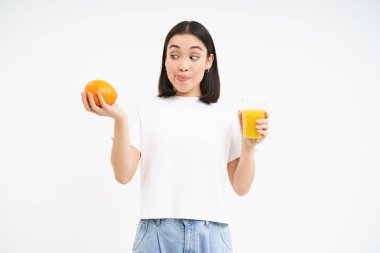 Tatlı Koreli kız, portakallı fuit ve meyve suyu arasında seçim yapıyor, her iki tarafa bakıyor, beyaz arkaplan.