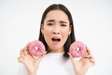 Üzgün Asyalı kadının portresi, diyette olduğu için üzgün, iki jöleli pembe çörek gösteriyor, abur cubur yemeye meyilli, beyaz arka plan.