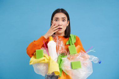 Şok olmuş Asyalı bir kadın, elinde plastik çöplerle, geri dönüşüm için atık ayıklarken, mavi arka planda korkunç bir şeyden utanıyor..