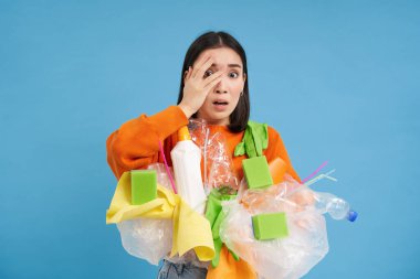 Şok olmuş Asyalı bir kadın, elinde plastik çöplerle, geri dönüşüm için atık ayıklarken, mavi arka planda korkunç bir şeyden utanıyor..
