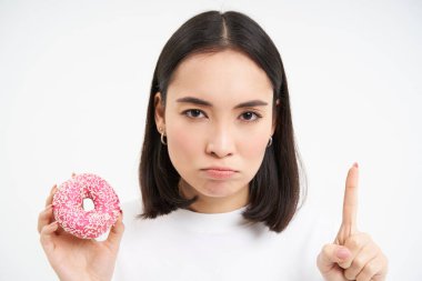 Ciddi Asyalı kadınlara yakın, tabu gösteriyor, jestleri ve pembe donutları durduruyor, şeker yemeyi yasaklıyor, beyaz arka plan.