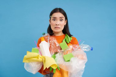 Farklı tipte plastikleri olan kafası karışmış genç Asyalı kız, geri dönüşüm yapmayı, çevreyi korumayı, çöpleri ayırmayı, mavi arka planı öğreniyor..
