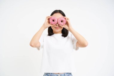 Tatlı gülümseyen Koreli kadın, donut deliklerinden bakıyor, lezzetli çörekler yiyor, beyaz arka planda izole edilmiş..