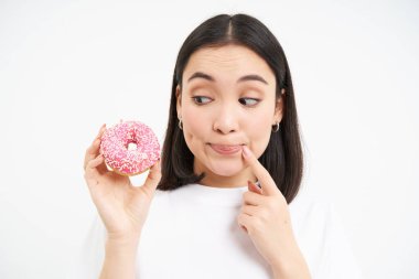 Gülümseyen genç Koreli kadın, düşünüyor ve bakıyor pembe jöleli donut, beyaz arka plan.