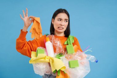Koreli kadın kokuşmuş lateks eldivene bakıyor, elinde plastik çöp tutuyor, evi ve geri dönüşümü temizliyor, mavi arka plan..