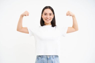 Asyalı genç bir kadının portresi, kaslarını kasıyor, kaslarını gösteriyor ve kendine güvenerek gülümsüyor, güçlü görünüyor, beyaz arka planda duruyor..