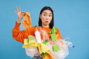 Genç Asyalı kadın elinde çöp, plastik şişeler ve atıklar tutuyor. Kirli lateks eldivene, mavi arka plana şaşırmış görünüyor..