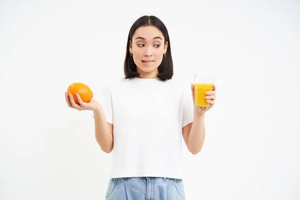 stock image Cute korean girl, makes choice between orange fuit and juice, looks both sides, white background.