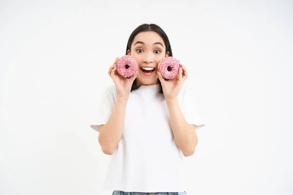 stock image Happy young asian woman, holding two glazed doughnuts near face and looking excited, likes eating dessert, junk food and takeaway concept.