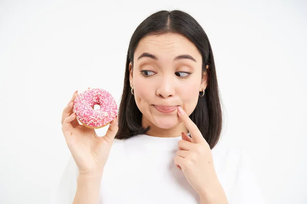 stock image Smiling young korean woman, thinking and gazing at pink glazed doughnut, white background.