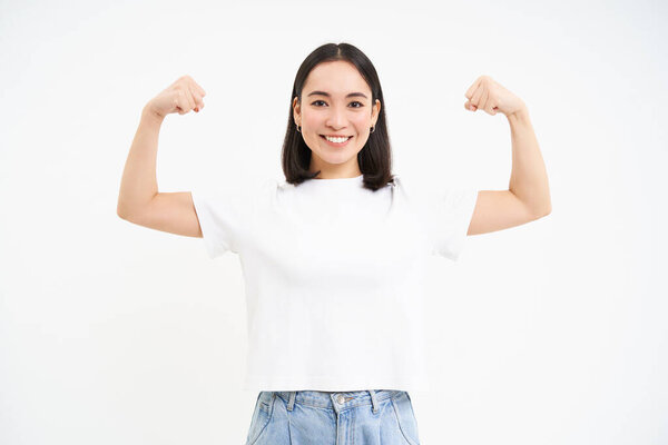 Портрет молодой азиатки, сгибающей мышцы, показывающей бицепсы и улыбающейся с уверенностью, выглядящей сильной, стоящей на белом фоне.
