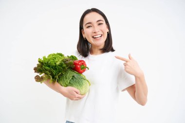 Mutlu Koreli kadın görüntüsü, sebzelere işaret eden, gülümseyen, sağlıklı çiğ yiyecekler yemeyi öneren, yeşil diyet kavramı, beyaz arka plan.