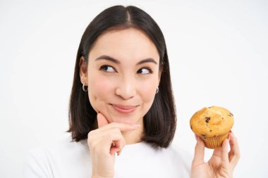 Düşünceli gülümseyen suratlı Asyalı bir kadının yakın portresi, elinde kek, hamur işi yemeyi düşünüyor, beyaz arka plan.