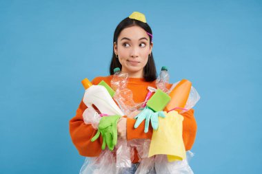 Kafası karışmış genç Asyalı kadın, elinde plastik şişeler, ev çöpleri, geri dönüşüm için atık ayıklama, mavi arka plan..