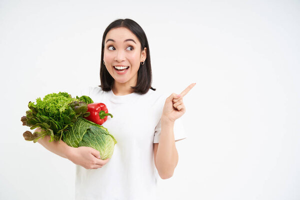 Enthusiastic korean woman, holding bunch of vegetables, pointing finger right at banner, showing promo advertisement, white background.