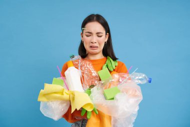 Üzgün Asyalı kız öğrenci, geri dönüşüm atıklarını tutuyor, geri dönüşüm merkezine götürmek için plastik çöp topluyor, mavi arka plan..