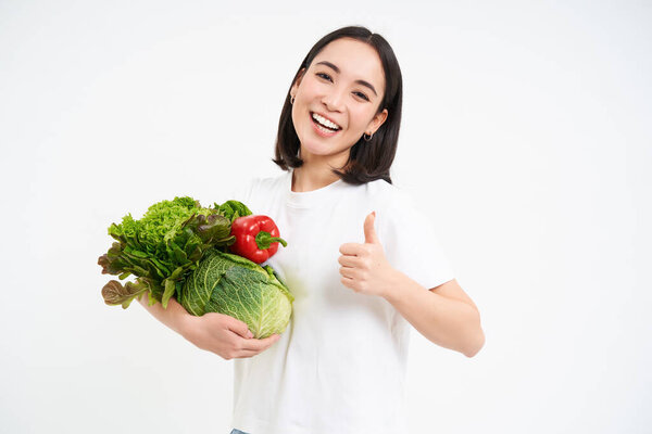 Smiling happy asian woman shows thumbs up and vegetables, eating healthy raw food, isolated over white background.