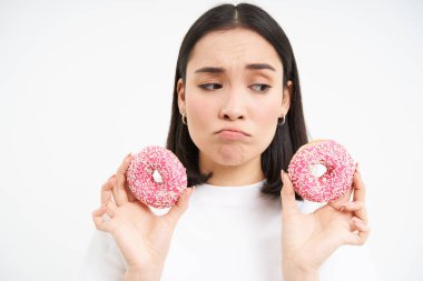 Üzgün Asyalı kadının portresi, diyette olduğu için üzgün, iki jöleli pembe çörek gösteriyor, abur cubur yemeye meyilli, beyaz arka plan.