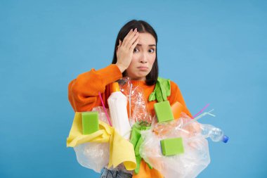 Şok olmuş Asyalı bir kadın, elinde plastik çöplerle, geri dönüşüm için atık ayıklarken, mavi arka planda korkunç bir şeyden utanıyor..