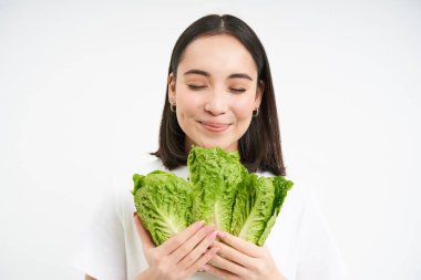 Gülümseyen Asyalı kadın, vejetaryen lahana yiyor, lezzetli taze marul yapraklarından hoşlanıyor, beyaz arka planda duruyor..