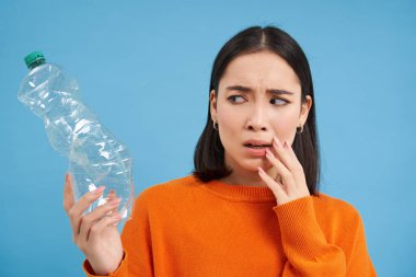 Endişeli Asyalı kadının portresi, plastik şişeye endişeli bakıyor, nerede geri dönüşüm yapacağını düşünüyor, mavi arka plan..