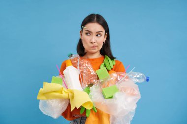 Eko kadın elinde plastik şişeler ve geri dönüşüm atıkları tutuyor, kalınlaşıyor, kafası karışmış görünüyor, çöpleri farklı kutulara ayırıyor, mavi arka plan..