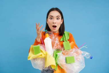 Asyalı kadın şok olmuş görünüyor, elinde plastik çöp tutuyor, geri dönüşüm için çöp ayırıyor, şaşırmış görünüyor, mavi arka plan..
