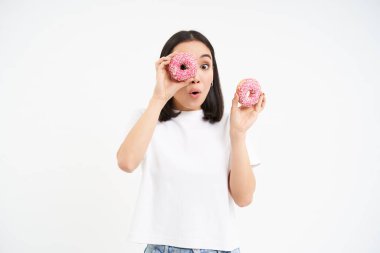 Tatlı gülümseyen Koreli kadın, donut deliklerinden bakıyor, lezzetli çörekler yiyor, beyaz arka planda izole edilmiş..