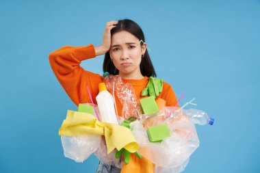Karmaşık yüzlü bir kadın, plastik çöplere kafası karışmış, eşyaları nasıl geri dönüştüreceğinden bihaber, yeşile dönmüş, mavi arka planda dikiliyor..