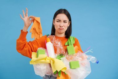 Koreli kadın kokuşmuş lateks eldivene bakıyor, elinde plastik çöp tutuyor, evi ve geri dönüşümü temizliyor, mavi arka plan..