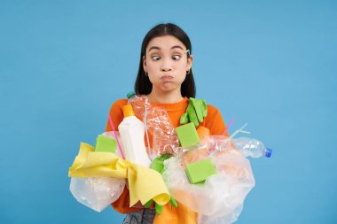 Genç kadın elinde plastik çöpler, geri dönüşümlü çöpler ve gözlerini kıs, geri dönüşüm kuralları ve mavi arka plan yüzünden kafası karışsın..