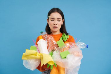 Üzgün Asyalı kız öğrenci, geri dönüşüm atıklarını tutuyor, geri dönüşüm merkezine götürmek için plastik çöp topluyor, mavi arka plan..