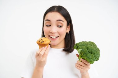 Asyalı kadın modellere yakın çekim, kek yemek, hamur işi yemek ve sağlıklı sebze brokolisine bakmak, beyaz arka plan..