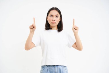 Şüpheci Asyalı kadın, şüpheli bir yüz ifadesiyle yukarı bakıyor, beyaz arka planda duruyor..