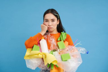 Sinirli genç bir kadın evi temizliyor, elinde geri dönüşüm için plastik çöp tutuyor, atık toplamaktan bıkmış, mavi arka planda dikiliyor..