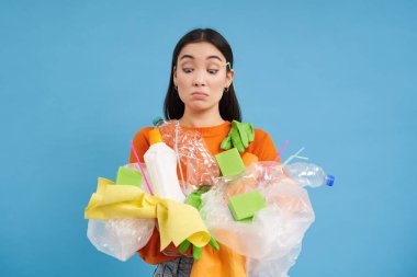 Asyalı kız ellerinde plastik atık, geri dönüşüm malzemeleri, geri dönüşüm için çöp toplama, mavi arka plan tutarken ellerine bakıyor..