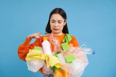 İğrenç Asyalı kadın, elinde kokuşmuş çöp tutuyor, atıklarını geri dönüştürüyor hayal kırıklığına uğramış, isteksiz yüz, mavi arka plan.