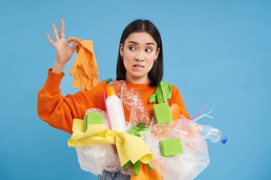 Genç kadın plastik çöpü tutuyor, şok olmuş yüzüyle eldivene bakıyor, pis kokulu atık, geri dönüşüm için çöp ayırıyor, mavi arka plan..